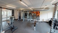 This fitness center is designed to accommodate a wide range of workout styles across a single, thoughtfully organized level. The space is divided into functional zones, beginning with a cardio area featuring multiple treadmills and elliptical machines positioned near large windows that allow natural light to fill the room and provide exterior views. A bold orange accent wall with geometric graphic elements serves as a striking visual focal point throughout the facility, adding energy and character to the environment.

The flooring transitions between a distinctive herringbone-patterned carpet in the cardio and entry areas and durable rubber flooring in the strength training zone, clearly delineating each area's purpose. The strength training section is well-appointed with cable machines, free weights, barbells, benches, and racks, lined by floor-to-ceiling mirrors that enhance the sense of space. Overhead, linear lighting illuminates exposed ductwork, lending an industrial aesthetic to this zone.

A rack of medicine balls and additional functional training equipment occupy a designated area near the accent wall. The facility also includes a vending area near the stairwell entry and offers views into an adjacent pool area through interior windows, rounding out the amenities available within this comprehensive fitness space.