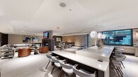 Located at 624 N Glebe Rd in Arlington, Virginia, this contemporary amenity lounge is a thoughtfully designed, single-floor space that seamlessly blends social and recreational functions. The open-plan layout flows effortlessly between distinct activity zones, creating a dynamic yet cohesive environment throughout.

At the heart of the space is a generous bar and counter area finished in polished white stone, lined with mid-century modern seating and illuminated by striking globe pendant lights. Adjacent to the bar, a fully equipped catering kitchen features a textured subway tile accent wall, dark stone countertops, wood-toned cabinetry, large windows overlooking an outdoor courtyard, and warm brass sconce lighting.

The lounge area offers multiple seating arrangements, including plush sectional sofas and leather accent chairs centered around a sleek double-sided fireplace with a stone surround and wall-mounted television. A dedicated game room includes a full-size billiards table and a shuffleboard table set against a distinctive textured wood slat accent wall, with direct access to the outdoors through glass doors.

A private screening or media lounge features deep upholstered bench seating, recessed lighting, and a large display screen. Throughout, light wood-tone flooring, recessed lighting, and a refined neutral palette contribute to a polished, welcoming ambiance ideal for both social gatherings and relaxed evenings.