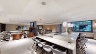 Located at 624 N Glebe Rd in Arlington, Virginia, this contemporary amenity lounge is a thoughtfully designed, single-floor space that seamlessly blends social and recreational functions. The open-plan layout flows effortlessly between distinct activity zones, creating a dynamic yet cohesive environment throughout.

At the heart of the space is a generous bar and counter area finished in polished white stone, lined with mid-century modern seating and illuminated by striking globe pendant lights. Adjacent to the bar, a fully equipped catering kitchen features a textured subway tile accent wall, dark stone countertops, wood-toned cabinetry, large windows overlooking an outdoor courtyard, and warm brass sconce lighting.

The lounge area offers multiple seating arrangements, including plush sectional sofas and leather accent chairs centered around a sleek double-sided fireplace with a stone surround and wall-mounted television. A dedicated game room includes a full-size billiards table and a shuffleboard table set against a distinctive textured wood slat accent wall, with direct access to the outdoors through glass doors.

A private screening or media lounge features deep upholstered bench seating, recessed lighting, and a large display screen. Throughout, light wood-tone flooring, recessed lighting, and a refined neutral palette contribute to a polished, welcoming ambiance ideal for both social gatherings and relaxed evenings.