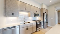 This thoughtfully designed single-floor unit showcases a clean, modern aesthetic throughout. The kitchen is a standout feature, outfitted with textured flat-panel cabinetry in a warm neutral tone, a herringbone-patterned tile backsplash, quartz countertops, and a full suite of stainless steel appliances including a gas range, built-in microwave, dishwasher, and French door refrigerator. A central island with a contrasting base provides additional prep and seating space, while sleek pendant lighting adds a refined touch overhead. Light hardwood flooring flows through the kitchen and into the open living area, contributing to a bright and airy atmosphere. A sliding glass door leads to a private balcony, offering views of the surrounding urban landscape and allowing abundant natural light to fill the space. The bedroom is finished with plush carpet and features large windows that frame outdoor views, creating a comfortable and well-lit retreat. The full bathroom is appointed with large-format marble-look tile flooring, a light wood vanity with a white quartz countertop, a framed mirror, and a tub-shower combination with clean white tile surround. Throughout the unit, neutral wall tones, black hardware accents, and consistent modern finishes create a cohesive and polished living environment.