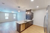 Located at Walnut St & Route 140 in Foxboro, MA, this thoughtfully designed one-bedroom, one-bathroom unit offers a functional and modern living experience across a single floor. The open-concept kitchen and living area serves as the heart of the home, anchored by a sleek center island with a white quartz countertop, undermount sink, and stainless steel dishwasher. Dark-toned cabinetry with contemporary hardware contrasts beautifully against the light countertops, while pendant lighting and recessed ceiling fixtures provide both task and ambient illumination. A stainless steel refrigerator and range with a ventilation hood complete the kitchen. Light hardwood flooring runs throughout, lending warmth and continuity to the space. The bedroom is a comfortable, well-proportioned retreat featuring multiple windows that invite abundant natural light and views of the surrounding wooded landscape. Two closets provide ample storage. The bathroom is designed with accessibility in mind, equipped with grab bars, a barrier-free floor, a dark-cabinet vanity with a mirror, and clean white walls. The unit's neutral color palette and clean-lined aesthetic create a bright, contemporary atmosphere throughout.
