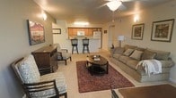 This single-floor unit showcases an open-concept layout that seamlessly connects the living area to the kitchen. The living room is comfortably appointed with a plush sofa, patterned accent chairs, a dark wood coffee table, and a rich burgundy area rug anchoring the space. A ceiling fan with a light fixture provides overhead illumination, complemented by wall-mounted sconce lighting that adds warmth to the neutral-toned walls and carpeted flooring. Framed artwork decorates the walls, contributing to a cozy, lived-in ambiance. The kitchen is positioned at the rear of the open space and features warm honey-toned cabinetry, white appliances including a refrigerator and microwave, and a raised breakfast bar with two bar stools — ideal for casual dining or entertaining. The open sightlines between the kitchen and living area enhance the sense of spaciousness throughout the unit. Overall, the space offers a functional and inviting layout with consistent warm, neutral finishes that create a cohesive and comfortable atmosphere.