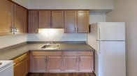 This single-floor unit presents a straightforward, functional layout with a neutral color palette throughout. The living areas are finished with wall-to-wall beige carpeting, smooth white walls, and flush-mount ceiling light fixtures, creating a clean and adaptable interior. Windows fitted with vertical blinds allow natural light to filter into the main rooms, contributing to a bright and open atmosphere.

The kitchen is equipped with warm wood-toned cabinetry spanning both upper and lower sections, providing ample storage. A stainless steel sink sits beneath under-cabinet lighting, complemented by speckled laminate countertops and wood-look flooring. A white refrigerator is included among the appliances.

The bedroom areas are generously sized, featuring the same plush carpeting found throughout the unit. Multiple closets are present, including what appears to be a walk-in closet, offering substantial storage capacity.

The entryway is accessed through a solid six-panel door and connects directly to a full bathroom, which includes a tiled tub-shower combination, a toilet, and tile flooring. The overall layout, as visible in the 3D model, is compact yet well-organized, with distinct zones for living, cooking, sleeping, and bathing, making practical use of the available space.