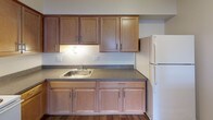 This single-floor unit presents a straightforward, functional layout with a neutral color palette throughout. The living areas are finished with wall-to-wall beige carpeting, smooth white walls, and flush-mount ceiling light fixtures, creating a clean and adaptable interior. Windows fitted with vertical blinds allow natural light to filter into the main rooms, contributing to a bright and open atmosphere.

The kitchen is equipped with warm wood-toned cabinetry spanning both upper and lower sections, providing ample storage. A stainless steel sink sits beneath under-cabinet lighting, complemented by speckled laminate countertops and wood-look flooring. A white refrigerator is included among the appliances.

The bedroom areas are generously sized, featuring the same plush carpeting found throughout the unit. Multiple closets are present, including what appears to be a walk-in closet, offering substantial storage capacity.

The entryway is accessed through a solid six-panel door and connects directly to a full bathroom, which includes a tiled tub-shower combination, a toilet, and tile flooring. The overall layout, as visible in the 3D model, is compact yet well-organized, with distinct zones for living, cooking, sleeping, and bathing, making practical use of the available space.