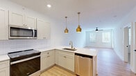 Located in Hendersonville, NC, this thoughtfully designed single-floor residence offers a clean, modern aesthetic with quality finishes throughout. The open-concept kitchen and living area serves as the heart of the home, featuring light hardwood-style flooring, recessed lighting, and a ceiling fan that keeps the space comfortable year-round. The kitchen is outfitted with crisp white shaker cabinetry accented by gold hardware, white quartz countertops, a subway tile backsplash, and a full suite of stainless steel appliances including a refrigerator, range, microwave, and dishwasher. A peninsula with an undermount sink doubles as a breakfast bar, illuminated by two striking gold pendant lights that add a warm, modern touch. The living area is bright and open, with natural light streaming through windows fitted with blinds and direct access via a glass-paneled door. The generously sized bedroom features soft carpeting, a ceiling fan, a window with outdoor views, and convenient closet access. The full bathroom is finished with light hardwood-style flooring, a white vanity, chrome fixtures, and a tub-shower combination. Clean white walls and consistent finishes throughout lend a cohesive, move-in-ready feel to this well-appointed Hendersonville residence.