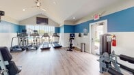 Located at 3434 Laurens Rd in Greenville, South Carolina, this single-floor fitness center is thoughtfully designed to support a well-rounded workout experience. The space features striking vaulted ceilings that lend a sense of openness and volume, while a bold two-tone color scheme — vivid blue on the upper walls transitioning to crisp white below — creates an energizing atmosphere throughout.

Large windows along the exterior walls flood the space with abundant natural light and offer pleasant views of the surrounding outdoor area, enhancing the workout environment. Light wood-style flooring runs throughout, contributing to a clean, modern aesthetic.

The cardio section is well-equipped with multiple treadmills, elliptical machines, and a stationary cycling bike, each positioned to take advantage of the natural light and outdoor views. A wall-mounted flat-screen television is prominently placed for entertainment during cardio sessions, with a ceiling fan providing additional airflow and comfort.

The strength training area is equally comprehensive, featuring a multi-function cable machine, a smith machine or power rack, adjustable benches, and a full rack stocked with kettlebells, medicine balls, and a complete set of dumbbells. TRX suspension training straps are also available for functional fitness workouts.

A direct exterior door provides convenient access to the outdoors, rounding out this fully equipped and inviting community fitness facility.