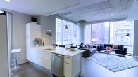 This single-floor space showcases a thoughtfully designed open-concept layout that seamlessly integrates the kitchen and living areas. The kitchen features sleek white cabinetry, a stainless steel undermount sink, and a dishwasher, all anchored by a central island with a clean white finish that provides additional workspace and casual seating. Pendant lighting with brushed metal fixtures hangs above the island, complementing the track lighting overhead.

The exposed concrete ceiling and structural column lend an industrial character to the space, while hardwood flooring adds warmth throughout. Floor-to-ceiling windows along two walls flood the interior with abundant natural light and frame dramatic panoramic views of the surrounding urban skyline, creating a strong connection to the city beyond.

The living area is open and generously proportioned, accommodating a full sofa arrangement atop a patterned area rug. The overall ambiance balances contemporary minimalism with industrial elements, resulting in a sophisticated, urban aesthetic suited for both everyday living and entertaining.
