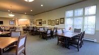This single-floor space centers around a well-appointed communal dining room designed to foster a warm and inviting atmosphere. The dining area is furnished with multiple dark-wood tables and cushioned chairs, arranged to accommodate both small and larger groups, making it well-suited for shared meals and social gatherings.

The walls are adorned with a curated collection of framed botanical and bird-themed artwork, lending the space a classic, refined character. A decorative wood trim border runs along the upper portion of the walls, adding architectural detail and visual continuity throughout the room. Fresh floral arrangements placed on select tables introduce subtle pops of color, enhancing the welcoming ambiance.

Natural light filters generously through large windows along one wall, brightening the carpeted interior and creating a pleasant, airy feel. The neutral-toned carpet and cream-colored walls contribute to a calm, comfortable environment. Recessed ceiling lighting supplements the natural light, ensuring consistent illumination throughout the day.

Adjacent to the main dining area, a service or kitchen space is visible in the background, suggesting convenient access to food preparation and service. Overall, the space is thoughtfully designed to prioritize comfort, community, and a dignified dining experience.