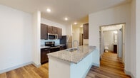 This single-floor unit showcases a clean, contemporary design with warm wood-look flooring that flows consistently throughout every room. The kitchen serves as a functional centerpiece, featuring a large peninsula island topped with speckled granite and an integrated sink. Rich dark-brown cabinetry lines the walls and pairs with a full suite of stainless steel appliances, including a refrigerator, electric range, and built-in microwave. Recessed lighting illuminates the space efficiently.

A hallway connects the kitchen to the remaining rooms, with a HVAC vent positioned overhead and a door providing access to additional spaces. The bedrooms are bright and well-proportioned, each equipped with ceiling fans and large windows fitted with horizontal blinds, allowing ample natural light and outdoor views. The neutral, cream-toned walls throughout contribute to an open and airy atmosphere.

The bathroom features a dark-wood vanity with a granite countertop, a framed mirror with bar lighting, and wood-look flooring. A separate shower area includes white subway-style tile walls, a chrome fixture set, and a clean white shower base. The overall design maintains a cohesive, modern aesthetic with consistent finishes and a practical layout throughout.