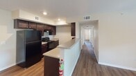 This single-story residence is characterized by consistent wood-style flooring that flows throughout every room, creating a cohesive and warm aesthetic. The kitchen is a functional galley-style space outfitted with rich dark espresso cabinetry extending to the upper walls, light-toned countertops, a double basin sink, and black appliances including a refrigerator and gas range. A peninsula counter separates the kitchen from the main living corridor, offering additional prep space and an open sightline toward the hallway leading to the bedrooms.

The living area benefits from natural light admitted through sizable windows fitted with horizontal blinds, complemented by a ceiling fan with integrated lighting. Multiple bedrooms follow a similar layout, each featuring ceiling fans, natural window views, and access to walk-in or reach-in closets with arched panel doors. One bedroom includes a conveniently located electrical panel access panel.

The bathroom presents a clean, practical design with a soaking tub and shower combination, dark wood-toned upper cabinetry for storage, a vanity with a light countertop, and matching wood-style flooring. Neutral beige wall tones throughout the unit contribute to a bright, understated ambiance well-suited for a variety of furnishing styles.