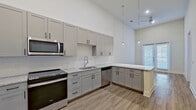 This thoughtfully designed single-floor residence offers a clean, contemporary aesthetic throughout. The kitchen serves as the centerpiece of the space, showcasing tall shaker-style cabinetry in a warm gray finish, granite countertops, a subway tile backsplash, and a full suite of stainless steel appliances including a French door refrigerator, electric range, over-the-range microwave, and dishwasher. A peninsula with additional cabinet storage provides extra counter space and opens naturally toward the living area. Pendant lighting drops elegantly from the soaring ceilings, enhancing the airy, open feel of the kitchen and adjoining space. Light wood-look flooring runs throughout the main living areas, complementing the neutral color palette. A glass-paneled exterior door allows natural light to pour in, brightening the space. The bedroom is generously sized, featuring plush carpeting, high ceilings, large windows with blinds, and direct access to a spacious walk-in closet outfitted with dual hanging rods and shelving on all three walls. The full bathroom is finished with large-format tile surrounding a soaking tub and shower, a granite-topped vanity with gray cabinetry, and wood-look flooring, creating a cohesive and polished look throughout the unit.