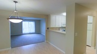This single-floor unit features a practical and open layout that connects the kitchen and living area seamlessly. The kitchen is equipped with white cabinetry, a built-in microwave, and a range, all finished in a clean, bright aesthetic. A peninsula-style pass-through counter separates the kitchen from the adjacent living space, creating an open and connected feel while maintaining a defined cooking zone. The flooring transitions from smooth laminate wood-look planks in the kitchen and entry area to plush carpeting in the living room, adding warmth and comfort underfoot. The living room is generously sized and benefits from a large window fitted with vertical blinds, allowing ample natural light to fill the space. Neutral beige tones on the walls throughout the unit contribute to a calm and versatile ambiance, making it easy to personalize with various décor styles. A textured ceiling adds a subtle design detail overhead. The overall layout is functional and straightforward, offering a comfortable living environment suitable for everyday use.