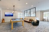 Located at 200 Travis Dr in McDonough, GA, this well-appointed single-floor community amenity space offers a thoughtfully designed layout ideal for socializing and recreation. The centerpiece is a spacious game room featuring a full-size pool table beneath a sleek linear chandelier, complemented by a mounted flat-screen TV and a comfortable lounge seating area with a contemporary sofa and accent chairs. A striking floor-to-ceiling black steel-framed glass partition wall serves as an architectural focal point, visually dividing the game and lounge zones while maintaining an open, connected feel throughout. Patterned carpet in muted blue-grey tones flows consistently across the space, adding texture and warmth underfoot. Natural light pours in through numerous large windows dressed with neutral drapery, creating a bright and welcoming atmosphere. A dedicated dining area features booth-style seating with upholstered benches and a wooden table, lit by modern pendant fixtures. A private meeting or dining room offers an intimate setting with a six-seat table, wall-mounted TV, and decorative wood slice wall art. An additional casual seating nook with a bistro table and chairs provides a quiet spot near sun-filled windows. A kitchen rounds out the amenities, supporting the space's versatile, community-focused function.