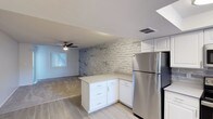This single-floor space offers a thoughtfully designed open-concept layout that seamlessly connects the kitchen and living areas. The kitchen is outfitted with crisp white cabinetry, light-toned countertops, and stainless steel appliances, including a full-size refrigerator. An L-shaped peninsula provides additional counter space and storage, making the kitchen both functional and visually appealing. A standout design feature is the textured faux brick accent wall in a neutral grey tone, which adds depth and character to the space while complementing the overall modern aesthetic. The flooring transitions from light wood-look planks in the kitchen to a neutral carpet in the living area, creating a natural visual boundary between the two zones. The living area is equipped with a ceiling fan, ensuring year-round comfort, and benefits from natural light streaming in through a window. A skylight above the kitchen further enhances the brightness of the space. The overall ambiance is clean, contemporary, and inviting, with a neutral color palette throughout that provides a versatile backdrop for a variety of interior styles.