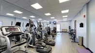 This fitness center is designed to support a comprehensive workout experience within a clean, functional layout. The space is lined with floor-to-ceiling mirrors along one wall, visually expanding the room and allowing users to monitor their form during exercise. Recessed panel lighting across the drop ceiling provides consistent, bright illumination throughout.

The cardio section is prominently featured, housing multiple elliptical trainers and additional aerobic machines arranged in rows, offering ample equipment for users to train simultaneously without feeling cramped. Toward the rear of the space, a cable and weight station is visible, extending the facility's offerings beyond cardio training. A rack of free weights or medicine balls is positioned along the far wall, rounding out the strength-training options.

The flooring is a wood-look laminate or vinyl material, practical and easy to maintain in a high-traffic fitness environment. Mounted flat-screen televisions provide entertainment for cardio users. Natural light filters in through a window near the back of the room, complementing the overhead lighting. The overall ambiance is bright, open, and purpose-driven, making it a practical and well-organized fitness facility.