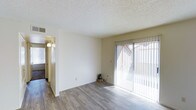 This single-floor unit offers a clean, well-lit living environment designed for comfortable everyday living. The main living area is an open, airy space characterized by smooth white walls and wood-look plank flooring that runs throughout, providing a modern yet warm aesthetic. A large sliding glass door fitted with vertical blinds serves as a prominent architectural feature, flooding the room with generous natural light while offering direct access to an outdoor area. The textured popcorn ceiling adds a classic residential touch consistent throughout the space. A hallway connects the living area to the rest of the unit, where wall-mounted lighting fixtures provide warm ambient illumination near the entryway. The neutral color palette of soft whites and beige tones creates a versatile backdrop suitable for a variety of interior styles. The overall layout is straightforward and functional, with an efficient use of space that maximizes openness. The combination of natural light streaming through the sliding door, the durable flooring, and the clean wall finishes gives the unit a bright, move-in-ready feel that is both practical and inviting.
