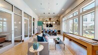 Located in Denton, Texas, this thoughtfully designed community clubhouse spans a generous single floor, offering a dynamic blend of social and functional spaces. The entry foyer greets visitors with soaring ceilings, large glass doors, and views of the outdoor pool area, setting an open and welcoming tone. Light-toned tile flooring and dramatic pendant lighting carry through the main living areas, which feature a sophisticated lounge with plush seating, arched architectural details, and floor-to-ceiling windows that flood the space with natural light. The kitchen and dining area stand out with bold teal cabinetry, striking botanical wallpaper, marble-veined countertops, warm wood open shelving, and a herringbone-patterned hardwood floor. A communal dining table with modern globe pendants anchors the space, complemented by a stainless steel refrigerator and full wet bar setup. A dedicated game room provides casual entertainment with a shuffleboard table and large windows overlooking the outdoors. Private office rooms, finished in rich teal paneling with wall-mounted displays and round work tables, offer quiet, focused workspaces. A marble-topped reception counter near the entrance completes the professional yet inviting atmosphere of this well-appointed clubhouse.