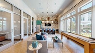 Located in Denton, Texas, this thoughtfully designed community clubhouse spans a generous single floor, offering a dynamic blend of social and functional spaces. The entry foyer greets visitors with soaring ceilings, large glass doors, and views of the outdoor pool area, setting an open and welcoming tone. Light-toned tile flooring and dramatic pendant lighting carry through the main living areas, which feature a sophisticated lounge with plush seating, arched architectural details, and floor-to-ceiling windows that flood the space with natural light. The kitchen and dining area stand out with bold teal cabinetry, striking botanical wallpaper, marble-veined countertops, warm wood open shelving, and a herringbone-patterned hardwood floor. A communal dining table with modern globe pendants anchors the space, complemented by a stainless steel refrigerator and full wet bar setup. A dedicated game room provides casual entertainment with a shuffleboard table and large windows overlooking the outdoors. Private office rooms, finished in rich teal paneling with wall-mounted displays and round work tables, offer quiet, focused workspaces. A marble-topped reception counter near the entrance completes the professional yet inviting atmosphere of this well-appointed clubhouse.