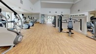 Located at 1699 E Washington St in Colton, California, this single-floor fitness center is a well-equipped facility designed to support a wide range of workout routines. The main gym area features soaring vaulted ceilings that create an open, airy atmosphere, with warm wood-look flooring throughout that adds a clean and inviting aesthetic. Large windows line the walls, allowing abundant natural light to fill the space while offering views of the outdoor pool area.

The cardio section is outfitted with multiple treadmills, elliptical machines, and a StairMaster, all thoughtfully arranged to maximize floor space. Strength training is well represented with a selection of cable machines, weight benches, and a full dumbbell rack. Full-length mirrors along one wall enhance both functionality and the sense of spaciousness in the main workout area.

A separate room dedicated to flexibility and functional training features light wood-style flooring and direct access to the outdoor pool patio through large glass doors. This room is stocked with stability balls in multiple sizes, resistance bands, medicine balls, and yoga mats, catering to stretching, balance, and low-impact exercise needs. The poolside views from this room add a bright, resort-like ambiance to every workout session.