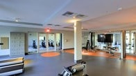 This single-floor fitness center offers a spacious and functional workout environment designed to accommodate a variety of exercise needs. The open layout provides ample room for movement, with a grey and orange-accented rubber flooring that adds a bold visual element while offering practical cushioning underfoot. A central support column subtly divides the space without disrupting the overall flow of the room.

The strength training area is equipped with a cable machine and adjustable bench, positioned alongside a large flat-screen television mounted on the wall. Mirrored walls line portions of the room, enhancing the sense of openness and allowing users to monitor their form during workouts. Cardio equipment, including stationary bikes and an elliptical, is distributed throughout the space.

Glass-paneled doors lead to an adjoining room visible through large mirrors, where additional cardio equipment such as upright bikes can be seen. The facility is well-lit with recessed ceiling lighting and benefits from natural light filtering through the glass entryway. Overhead HVAC vents ensure proper climate control throughout the space, contributing to a comfortable and functional fitness environment.