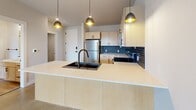 Located at 155 E 4th Place in Sioux Falls, SD, this modern one-bedroom, one-bathroom residence offers a thoughtfully designed open-concept layout across a single floor. The combined kitchen and living area serves as the heart of the home, showcasing light maple cabinetry, a striking navy blue subway tile backsplash, stainless steel appliances, and a peninsula with a matte black double sink and pendant lighting overhead. Polished concrete flooring runs throughout the main living areas, lending an industrial-modern aesthetic that ties the space together. The living room is filled with natural light and opens directly onto a private balcony through large black-framed sliding glass doors, offering views of the Sioux Falls cityscape. A ceiling fan adds comfort to the airy living space. The bedroom features a stylish barn door entry, a large black-framed grid window with additional city views, a ceiling fan, and a dedicated closet. The bathroom is finished with wood-look flooring, a white tub-shower combo, and matte black fixtures and hardware that echo the design language throughout the unit. A utility room provides added convenience. Clean lines, contemporary finishes, and abundant natural light define this well-appointed urban residence.