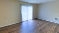 This single-story space presents a straightforward and functional layout highlighted by rich, dark hardwood flooring that extends throughout the main living area. The flooring adds warmth and a sense of depth to the room, complementing the neutral, light beige walls that create a clean and welcoming atmosphere.

A prominent sliding glass door serves as the focal point of the main living area, fitted with vertical blinds that allow for flexible light control. When open, the blinds permit generous amounts of natural light to filter into the space, brightening the interior and creating an airy feel. The door also provides direct access to an outdoor area, visible through the glass.

The textured ceiling adds a subtle architectural detail consistent with classic residential construction. White baseboards provide a crisp contrast against both the walls and flooring, contributing to the overall tidy appearance of the space.

The room is notably open and versatile, offering ample floor space that can accommodate a variety of furniture arrangements. The overall ambiance is calm and neutral, providing a blank canvas suitable for a range of interior styles and personal preferences.