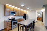 Located in Parkhaus in Lincoln, Nebraska, this single-floor unit offers four bedrooms, two bathrooms, a dedicated closet, and a combined kitchen and living area. The kitchen is well-appointed with warm wood cabinetry extending to the ceiling, stainless steel appliances including a gas range, microwave, dishwasher, and refrigerator, complemented by a tile backsplash and light tile flooring. A high-top dining table with industrial-style bar stools anchors the kitchen area, with stylish geometric light fixtures overhead adding a modern touch. Each of the four bedrooms features neutral-toned carpet flooring, crisp white walls, and individual windows fitted with blinds that frame views of the surrounding urban landscape. The primary bedroom includes both a window and a glass door leading to a private balcony, enhancing its sense of space and connection to the outdoors. One bathroom features a bathtub with accessible grab bars and tile flooring, while the second includes a vanity with wood cabinetry and a granite-style countertop, both finished with light tile floors. Clean lines, neutral tones, and functional layouts define the overall aesthetic of this practical yet comfortable multi-bedroom unit.