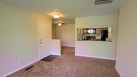 This single-floor residence offers a practical and comfortable open-concept layout designed for everyday living. The main living area is finished with wall-to-wall carpeting, creating a warm and cozy atmosphere throughout. A ceiling fan with an integrated light fixture provides both airflow and ambient lighting, complementing the flush-mount light near the entryway.

The front entry features a classic paneled white door with a dark threshold mat, providing a clean transition into the living space. A half-wall partition subtly defines the boundary between the entryway and the main living area, adding architectural interest without closing off the space.

The kitchen is positioned adjacent to the living area and is accessible via a pass-through opening in the shared wall, allowing for easy interaction between the two spaces. Light wood cabinetry lines the kitchen walls, paired with dark countertops that provide a grounding contrast. A black refrigerator and additional appliances are visible, indicating a fully equipped kitchen setup. A mounted microwave or television is also present above the counter area, adding functionality.

The walls throughout are painted in a neutral, soft green-beige tone, contributing to a calm and understated ambiance. Overall, this single-floor layout is thoughtfully arranged for comfortable, day-to-day living.