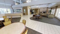 This single-floor community clubhouse offers an expansive, open-plan interior designed for comfort and social gathering. The main hall is immediately distinguished by its soaring high ceilings with recessed upper levels, elegant chandelier lighting, and large floor-to-ceiling glass doors and windows that flood the space with natural light and provide views of the surrounding outdoor area, complete with visible patio umbrellas and lush landscaping.

A centerpiece grand piano anchors the main gathering area, complemented by comfortable seating arrangements, carpeted lounge zones, and tile flooring that defines the traffic and activity areas. Decorative columns add an architectural formality to the space, while an American flag display adds a patriotic touch near the entrance.

The open layout extends into adjacent areas featuring a reception or service counter, additional seating and dining tables with upholstered chairs, and a cozy lounge with sofas and a fireplace. Warm earth-tone walls and neutral furnishings throughout create an inviting, relaxed ambiance. Multiple doorways lead to secondary rooms, offering functional spaces for a variety of community activities. The seamless flow between areas and the abundance of natural light make this clubhouse a bright, versatile, and welcoming gathering place.