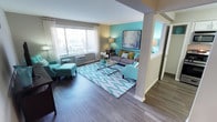 This single-floor residence centers around a welcoming living area that immediately draws attention with its bold teal accent wall, complemented by neutral tones throughout the rest of the space. The room is anchored by rich, dark hardwood-style flooring that runs seamlessly underfoot, adding warmth and contrast to the vibrant color palette. A large picture window with sheer white curtains floods the space with natural light, offering a bright and airy atmosphere while framing views of the exterior. A wall-mounted air conditioning unit sits beneath the window, ensuring year-round comfort. The living area is generously proportioned, accommodating a full seating arrangement that includes a grey sofa accented with decorative pillows, a teal accent chair, and a matching armchair with ottoman. A glass-top coffee table with a metallic frame rests atop a patterned area rug in coordinating teal and white tones, tying the design together. Artwork above the sofa echoes the room's color scheme. A structural column near the room's center subtly defines the space. An adjacent hallway, partially visible, suggests additional rooms beyond.