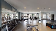 The Carriages on Lindberg Fitness Center is a spacious, single-floor facility designed to accommodate a comprehensive range of workout needs. The interior features light blue-gray walls, recessed lighting, and ceiling fans that contribute to a clean, comfortable environment. Large windows positioned throughout the space allow natural light to flow in, enhancing the overall atmosphere.

The cardio zone is well-appointed with multiple treadmills, elliptical machines, a stair climber, and a stationary spin bike, each positioned to face wall-mounted flat-screen televisions. A rowing machine is also available for low-impact, full-body training. Floor-to-ceiling mirrors line portions of the walls, visually expanding the space and supporting form-checking during exercise.

The strength training area includes a full rack of dumbbells spanning a wide weight range, multiple adjustable benches, a cable machine, a functional trainer, and a power rack. Rubber flooring in designated lifting zones provides cushioning and durability.

A dedicated stretching and open floor area features interlocking mats, exercise balls, and plyometric boxes for functional training. The entry vestibule includes a water bottle filling station, waste bin, and posted fitness center guidelines. Multiple access points and a clean, organized layout make this facility easy to navigate.