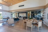 Located at 202 Mosley Ave in Alameda, California, this single-floor community clubhouse is a thoughtfully designed gathering space that combines modern aesthetics with a welcoming, resort-inspired atmosphere. The interior is immediately striking for its soaring vaulted ceilings finished in warm, natural wood planking, which lend a sense of openness and organic warmth throughout the entire space.

Large floor-to-ceiling windows and glass double doors flood the interior with abundant natural light while offering unobstructed views of the surrounding outdoor areas and lush greenery. The flooring transitions between large-format neutral tiles and light hardwood, reinforcing the clean, contemporary palette.

The main lounge area is anchored by a dramatic floor-to-ceiling textured slate accent wall housing a mounted flat-screen TV above a modern fireplace, creating a cozy focal point surrounded by generous sectional seating. A dedicated dining area with a round table and woven chairs sits adjacent, separated by glass-panel sliding doors that allow for flexible use of the space.

A secondary seating area provides a more intimate setting with armchairs, accent stools, and a stylish velvet sofa arranged around a decorative feature wall displaying curated artwork. Lush potted plants are distributed throughout, adding vibrant greenery that complements the bright, airy character of this well-appointed community space.