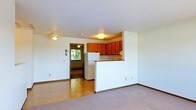 This single-floor residential space offers a practical and functional layout centered around an open-concept design. The living area is finished with plush carpet underfoot and smooth, neutral-toned walls that create a clean, understated backdrop. A textured ceiling with flush-mount lighting provides even illumination throughout the space.

The kitchen is positioned along one wall and is defined by a half-wall peninsula that separates it from the living area, offering a casual sense of openness between the two spaces. Warm, medium-toned wood cabinetry lines the upper section of the kitchen, providing ample storage, while a full-size refrigerator anchors the setup. The kitchen floor transitions to hardwood-style flooring, adding a visual distinction from the carpeted living zone.

Adjacent to the main living area, a connected room is accessible through a doorway and benefits from natural light streaming in through a window with a view of greenery outside. This additional room features the same hardwood-style flooring, giving it a cohesive yet distinct character. Overall, the space is straightforward and well-organized, with a layout that promotes ease of movement and a comfortable, livable atmosphere suited to everyday use.