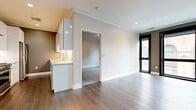 Located at 19 North St in Buffalo, NY, this thoughtfully designed single-floor space combines modern aesthetics with high-quality finishes throughout. The open living area is anchored by wide-plank hardwood flooring in a warm gray tone, recessed lighting, and crisp crown molding that frames the clean, neutral walls. A striking wall of floor-to-ceiling black-framed windows floods the main living space with natural light while offering views of Buffalo's historic neighboring architecture.

The kitchen is a standout feature, outfitted with sleek, high-gloss white cabinetry extending to the ceiling, stone countertops, a stainless steel range with a professional hood, and a full-size stainless steel refrigerator. A secondary prep sink with under-cabinet lighting adds additional functionality to this well-appointed space.

The carpeted bedroom offers a quieter, comfortable retreat, with its own black-framed window that frames views of the surrounding historic streetscape. Additional rooms provide flexible living arrangements across the single floor.

The bathroom is elegantly finished with marble-look tile covering both the walls and the walk-in shower surround, complemented by hexagonal floor tile, a wood-toned vanity with open shelving, a stone countertop, and a backlit mirror — delivering a spa-like atmosphere with refined, modern detailing throughout.
