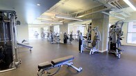 This fitness center offers a spacious, well-organized workout environment designed to accommodate a wide range of exercise needs. The space features an industrial aesthetic, highlighted by an exposed ceiling with visible ductwork and piping, painted in a neutral tone that complements the concrete-textured walls. Overhead, linear LED lighting fixtures provide consistent, bright illumination throughout the floor.

The gym is outfitted with an impressive selection of strength training machines, including cable systems, multi-function units, and a weight bench positioned prominently in the foreground. Cardio equipment, such as ellipticals and upright bikes, is visible toward the rear of the space. Large windows along the perimeter walls allow natural light to filter in, enhancing the open and airy feel of the room.

Dark rubber flooring covers the entire workout area, providing durability and safety underfoot. Full-length mirrors are mounted along the walls, serving both a functional and visual purpose by amplifying the sense of space. The overall layout is thoughtfully arranged to allow ample room between equipment, promoting ease of movement and a comfortable workout experience.