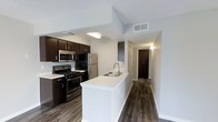 This single-floor residence offers a thoughtfully designed open-concept layout that seamlessly connects the kitchen and living areas. The kitchen is a standout feature, showcasing rich dark espresso cabinetry that contrasts beautifully with crisp white countertops. Stainless steel appliances — including a gas range, microwave, and refrigerator — provide a sleek, modern aesthetic. A peninsula island with an integrated double sink adds both functionality and a natural gathering point, making the kitchen ideal for cooking and entertaining alike.

Dark hardwood-style flooring runs throughout the space, adding warmth and visual continuity from the kitchen through the hallway. The neutral white walls and textured ceiling keep the interior feeling bright and clean, while recessed HVAC vents contribute to a streamlined look. A wide hallway leads further into the residence, hinting at additional rooms beyond, including what appears to be a spacious closet or storage area fitted with dark double doors. The overall ambiance is contemporary and well-appointed, balancing practical design elements with a cohesive, modern aesthetic suited for comfortable everyday living.