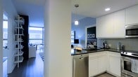 This single-floor residence showcases a thoughtfully designed open-concept layout that seamlessly connects the kitchen and living areas. The kitchen is appointed with crisp white cabinetry, dark granite countertops, and stainless steel appliances, including a dishwasher. Pendant and recessed lighting illuminate the space, adding both function and ambiance. A structural column subtly defines the transition between the kitchen and the living area without interrupting the open flow. The living area benefits from floor-to-ceiling windows that flood the space with natural light and provide expansive views of the surrounding cityscape. A black-and-white striped area rug anchors the living space, complementing the light hardwood flooring. The bedroom area, visible through the open layout, features a distinctive accent wall with a subtle patterned wallpaper in soft blue tones, adding a touch of personality and depth. Framed artwork adorns the walls, contributing to the curated, modern aesthetic. The overall design balances clean, contemporary lines with warm, inviting details, creating a residence that is both stylish and functional.