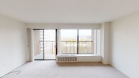 This well-appointed single-floor residence offers a clean, modern aesthetic throughout its thoughtfully designed layout. The generous living area is carpeted in neutral tones and bathed in natural light courtesy of expansive floor-to-ceiling windows and a sliding glass door that opens directly onto a private balcony. From this outdoor space, residents enjoy impressive views of the surrounding cityscape and distant mountain ridgeline, lending a scenic quality to everyday living. The interior entry area features durable hardwood-style flooring that transitions smoothly into the adjoining kitchen, keeping the spaces visually distinct. The kitchen is outfitted with rich, dark cabinetry contrasted by light stone-finish countertops, and is equipped with stainless steel appliances including a dishwasher, electric range, and built-in microwave. A single-basin sink and ample upper and lower cabinet storage complete this functional culinary space. The bathroom maintains the home's clean, contemporary palette with a white vanity, dark cabinetry base, and a shower-tub combination with sleek chrome fixtures. Crisp white walls and smooth ceilings throughout contribute to a bright, airy ambiance that makes the space feel open and inviting.
