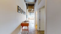 This 1-bedroom, 1-bathroom loft showcases a compelling industrial aesthetic rooted in its architectural bones. Soaring ceilings with exposed concrete, visible ductwork, and structural beams set the tone throughout, while polished concrete floors unify the single-level layout. A central hallway connects the living areas to the bedroom and bathroom, with exposed ceiling infrastructure and linear lighting running its length, adding to the loft's raw character.

The open-plan kitchen is a standout feature, anchored by a large island with a white quartz countertop and seating for two. Dark lower cabinetry contrasts with white upper cabinets, and a white subway tile backsplash frames the full suite of stainless steel appliances, including a French door refrigerator, range, and microwave. Pendant lights with bold black-and-yellow shades hang overhead, adding a modern accent.

The living area benefits from an expansive bank of steel-framed factory windows that span the far wall, delivering generous natural light and open views. An original concrete column with a teal accent detail adds a distinctive architectural touch. The bedroom is a private, well-proportioned retreat with clean white walls. Overall, the space blends urban industrial character with modern, functional finishes.