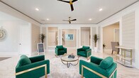 This thoughtfully designed single-floor space offers a refined and welcoming atmosphere, highlighted by gleaming large-format marble-look tile flooring that flows seamlessly throughout. The main living area commands attention with its open layout, framed by white decorative columns and wainscoting that lend a classic architectural character to the warm, neutral-toned walls. Dual ceiling fans with gold hardware complement the recessed lighting, ensuring the space remains bright and comfortable. Striking emerald green velvet armchairs with gold-trimmed frames are arranged around a glass-top coffee table, creating an inviting and stylish focal point. A shiplap accent wall adds texture near a built-in refreshment station, while tall tropical potted plants introduce a natural, organic element throughout. The hallway connects seamlessly to the dining room, which features a round white table surrounded by rattan chairs, flanked by large windows dressed with white blinds that invite views of lush tropical greenery outside. A wall-mounted display screen and globe pendant floor lamps complete the dining area's relaxed yet polished ambiance. Large windows along the perimeter bathe the entire space in natural light, enhancing the airy, upscale feel of this beautifully appointed interior.