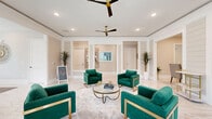 This thoughtfully designed single-floor space offers a refined and welcoming atmosphere, highlighted by gleaming large-format marble-look tile flooring that flows seamlessly throughout. The main living area commands attention with its open layout, framed by white decorative columns and wainscoting that lend a classic architectural character to the warm, neutral-toned walls. Dual ceiling fans with gold hardware complement the recessed lighting, ensuring the space remains bright and comfortable. Striking emerald green velvet armchairs with gold-trimmed frames are arranged around a glass-top coffee table, creating an inviting and stylish focal point. A shiplap accent wall adds texture near a built-in refreshment station, while tall tropical potted plants introduce a natural, organic element throughout. The hallway connects seamlessly to the dining room, which features a round white table surrounded by rattan chairs, flanked by large windows dressed with white blinds that invite views of lush tropical greenery outside. A wall-mounted display screen and globe pendant floor lamps complete the dining area's relaxed yet polished ambiance. Large windows along the perimeter bathe the entire space in natural light, enhancing the airy, upscale feel of this beautifully appointed interior.