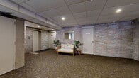 Located at 2727 N Maryland Ave in Milwaukee, Wisconsin, this ground-floor common area serves as the primary lobby and resident services hub for the building. The space is defined by its distinctive exposed brick accent walls, which lend a warm, textural character throughout. Underfoot, durable patterned carpet in earthy tones extends across the entire floor, providing both comfort and practicality in a high-traffic environment.

The lobby is accessed through a striking glass curtain wall entryway with dark metal framing, offering a transparent view from the interior to the building's exterior vestibule, where mailboxes line the walls on either side. Recessed lighting in the drop tile ceiling provides consistent, even illumination throughout the space.

A welcoming seating area features a cushioned settee and an armchair, accented with potted fiddle-leaf fig and tropical plants that add a touch of greenery. A framed mirror on the brick wall enhances the sense of openness. Adjacent to the seating area, a row of dark wood cabinetry serves as an organized package sorting and storage station, labeled by floor for resident convenience. A separate alcove with textured walls houses an additional seating nook beside a labeled utility room. An elevator provides vertical access within the building.