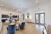 Located at 560 Jack Clift Blvd in Madison, Alabama, this beautifully designed single-floor community space offers an open-concept layout that seamlessly connects the living room, dining area, and kitchen. Soaring ceilings throughout create an airy, expansive atmosphere, while light wood flooring unifies the space with a warm, contemporary feel.

The living area is anchored by a striking multi-arm chandelier and features elegant furnishings, including a neutral sofa and accent chairs, alongside a decorative credenza topped with an arched mirror. Glass-panel double doors open directly to an outdoor patio, flooding the room with natural light and providing views of the surrounding community.

The kitchen is a standout feature, equipped with dark wood cabinetry, a large white quartz island with bar seating, and bold globe pendant lights with brushed gold finishes. The open layout transitions naturally into the dining area, which is bathed in natural light from oversized windows offering views of the landscaped grounds.

A separate reception or workspace area features light wood built-in cabinetry, pendant lighting, and an open, welcoming design. Throughout the space, crisp white walls, recessed lighting, and carefully curated décor contribute to a polished, modern ambiance well-suited for community gatherings and daily use.
