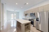 This well-designed 1-bedroom, 1-bathroom residence offers a thoughtful layout across a single floor, combining modern aesthetics with practical functionality. The open-concept kitchen and living area serves as the heart of the home, featuring light wood-grain cabinetry, a subway tile backsplash, stainless steel appliances including a refrigerator with ice maker, electric range, and built-in microwave. A central kitchen island with a clean white countertop provides additional prep space and casual seating. Light hardwood-style flooring flows throughout the main living areas, complementing the bright, neutral color palette. The living room benefits from natural light streaming through double doors that open to an exterior view, and a ceiling fan adds comfort overhead. The bedroom is a comfortable, carpet-floored retreat with a large window that invites natural light and outdoor views. The bathroom is well-appointed with a soaking tub and tiled shower surround, a vanity with storage, and access to a walk-in closet with built-in shelving. A dedicated laundry room with a full-size washer and dryer adds everyday convenience. Recessed lighting throughout the unit enhances the clean, contemporary ambiance.