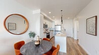 This contemporary single-floor residence showcases a thoughtfully designed open-concept layout that seamlessly connects the kitchen, dining, and living areas. The kitchen is a standout feature, outfitted with floor-to-ceiling white cabinetry, a herringbone tile backsplash, stainless steel appliances including a French door refrigerator and range, and a sleek white island with an undermount sink and pendant lighting above. Warm hardwood-style flooring runs consistently throughout the space, complementing the clean white walls and recessed lighting that keep the interiors bright and inviting. The dining area accommodates a round dark-wood table with terracotta velvet chairs, while a round wood-framed mirror adds a decorative accent. The living room offers an airy, relaxed atmosphere with natural light streaming through large sliding glass doors that open to a private balcony with urban views of the surrounding cityscape. The bedroom is a serene retreat featuring its own balcony access, a large window, and ample natural light. The bathroom maintains the residence's modern aesthetic with white cabinetry, a undermount sink, a wide frameless mirror, and hardwood-style flooring. Throughout, the design prioritizes clean lines, neutral tones, and functional elegance.