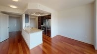 This single-floor residence offers a thoughtfully designed layout that balances open living areas with functional private spaces. Rich hardwood flooring extends throughout the main living and kitchen areas, adding warmth to the bright, neutral interiors. The kitchen is well-equipped, featuring dark wood cabinetry, stainless steel appliances including a refrigerator, gas range, and microwave, and a peninsula with a granite-style countertop and undermount sink — seamlessly connecting to the open living space. Natural light pours in through large windows and a full-width glass sliding door, leading to a private balcony that offers elevated views of the surrounding urban and wooded landscape. The bedroom is carpeted for comfort and includes floor-to-ceiling sliding glass access to the same balcony, further enhancing the sense of openness. The bathroom is clean and modern, finished in white with tile flooring, a walk-in shower with built-in shelving, a single-sink vanity with a white countertop, and an illuminated mirror. The entry area features hardwood flooring, a glass-paneled front door, and clean white walls that set a bright, welcoming tone throughout the unit.