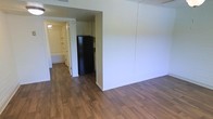 This single-floor unit offers a straightforward, functional layout designed to maximize usable space. The main living area features warm wood-look laminate flooring that extends seamlessly throughout, creating a cohesive and easy-to-maintain surface underfoot. Neutral white walls keep the space feeling clean and open, providing a blank canvas for personalization.

A partial wall divides the living area from the compact kitchen, which is equipped with a black refrigerator and overhead cabinetry. The kitchen's efficient design makes good use of its footprint while remaining accessible from the main living space.

A hallway visible from the living area leads to additional rooms, including what appears to be a bathroom and a space featuring built-in shelving — ideal for storage or use as a closet area. The hallway is well-lit, contributing to the overall brightness of the unit.

The layout flows naturally from the living area through to the back of the unit, offering a practical and organized arrangement. The consistent flooring and neutral color palette throughout give the space a unified, tidy appearance.
