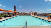 This amenity space offers an impressive combination of outdoor and indoor features designed for comfort and leisure. The centerpiece is a generously sized rooftop swimming pool with vivid turquoise water, framed by a spacious concrete deck lined with blue and orange lounge chairs and coordinating shade umbrellas. The rooftop setting provides breathtaking panoramic views of the Atlanta skyline, with iconic high-rise towers visible against open blue skies, creating a striking urban backdrop. Vibrant potted planters add pops of greenery and color throughout the deck.

Indoors, the lounge area is thoughtfully designed with a neutral palette of warm grey tones, light wood-look flooring, and a textured tile accent wall. The space accommodates both collaborative and casual seating, including a substantial dark wood communal table with bar stools and a pair of contemporary blue accent chairs positioned near large windows that bring in ample natural light and garden views. Industrial-style flush-mount fixtures illuminate the space from above.

Adjacent to the lounge is a dedicated coffee station featuring rich dark wood cabinetry, granite countertops, open wood shelving, and Starbucks-branded equipment — providing a café-quality refreshment experience. A pass-through window connects the lounge to an adjoining area, enhancing the open, connected feel of the space.