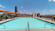 This amenity space offers an impressive combination of outdoor and indoor features designed for comfort and leisure. The centerpiece is a generously sized rooftop swimming pool with vivid turquoise water, framed by a spacious concrete deck lined with blue and orange lounge chairs and coordinating shade umbrellas. The rooftop setting provides breathtaking panoramic views of the Atlanta skyline, with iconic high-rise towers visible against open blue skies, creating a striking urban backdrop. Vibrant potted planters add pops of greenery and color throughout the deck.

Indoors, the lounge area is thoughtfully designed with a neutral palette of warm grey tones, light wood-look flooring, and a textured tile accent wall. The space accommodates both collaborative and casual seating, including a substantial dark wood communal table with bar stools and a pair of contemporary blue accent chairs positioned near large windows that bring in ample natural light and garden views. Industrial-style flush-mount fixtures illuminate the space from above.

Adjacent to the lounge is a dedicated coffee station featuring rich dark wood cabinetry, granite countertops, open wood shelving, and Starbucks-branded equipment — providing a café-quality refreshment experience. A pass-through window connects the lounge to an adjoining area, enhancing the open, connected feel of the space.