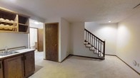 This two-story home presents a straightforward and functional layout with neutral interiors throughout. The main level features wall-to-wall carpeting in a warm beige tone, providing a soft and comfortable underfoot feel across the living areas. A prominent wood-trimmed staircase with dark wooden balusters connects the two floors, serving as a central architectural element on the main level. The staircase is positioned openly within the space, contributing to an unobstructed flow between rooms.

Dark wood interior doors with brass hardware are consistent throughout, complementing the wood baseboards and trim that frame each room. The ceilings feature a textured finish, adding subtle dimension to the otherwise clean white surfaces. Recessed and flush-mount lighting provides interior illumination throughout the space.

A glimpse into an adjacent room reveals cabinetry and fixtures consistent with a bathroom, suggesting practical amenities are conveniently located on the main floor. The overall color palette is neutral, with white walls and light carpet tones creating a bright and easy-to-customize interior. The home's design reflects a classic, no-frills residential style suited for comfortable everyday living.